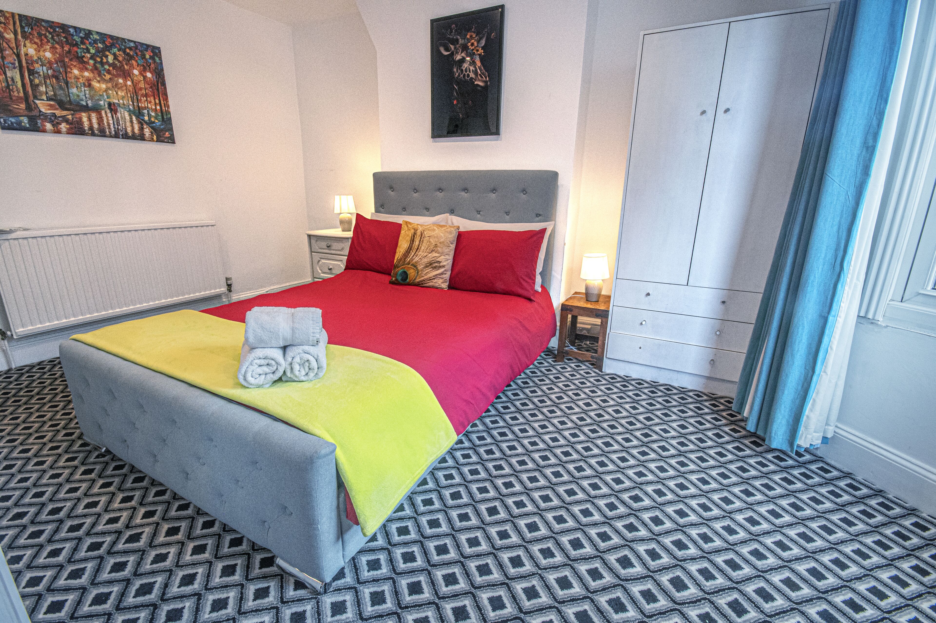 A neatly made bed with a red and yellow blanket, a grey upholstered headboard, and a patterned floor.