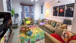 Ein Wohnzimmer mit einer Couch, einem Couchtisch und einem farbenfrohen Teppich. Es gibt ein Fenster mit Jalousien, eine Pflanze und eine Lampe auf einem Beistelltisch.