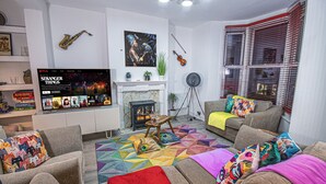 Ein Wohnzimmer mit Kamin, Sofas, bunten Kissen, einem Fernseher, der 'Stranger Things' zeigt, und einem Saxophon an der Wand.