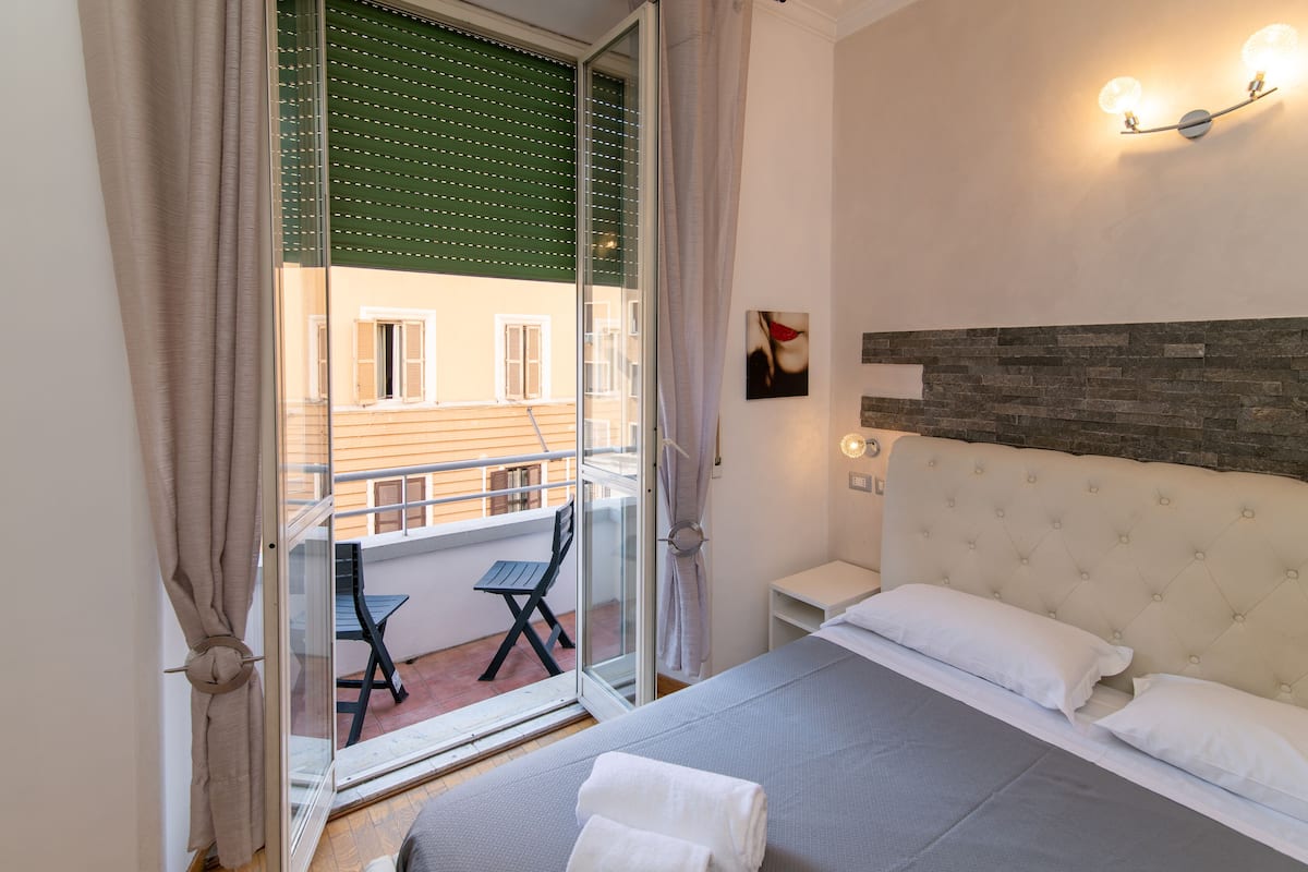 A bedroom with a bed, a nightstand, a wall-mounted light fixture, and a view of a balcony with a table and chairs.