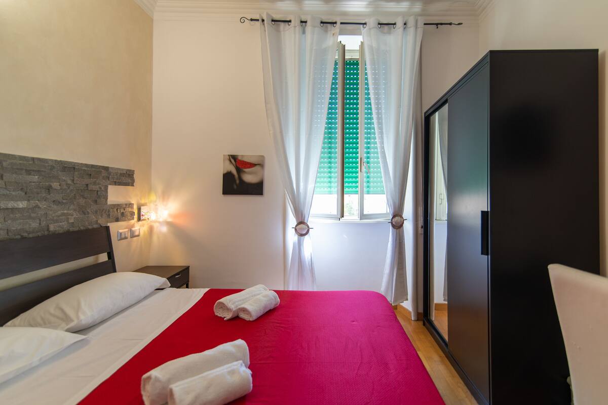 A hotel room with a bed, a nightstand, a wall-mounted light, a window with curtains, and a wardrobe.