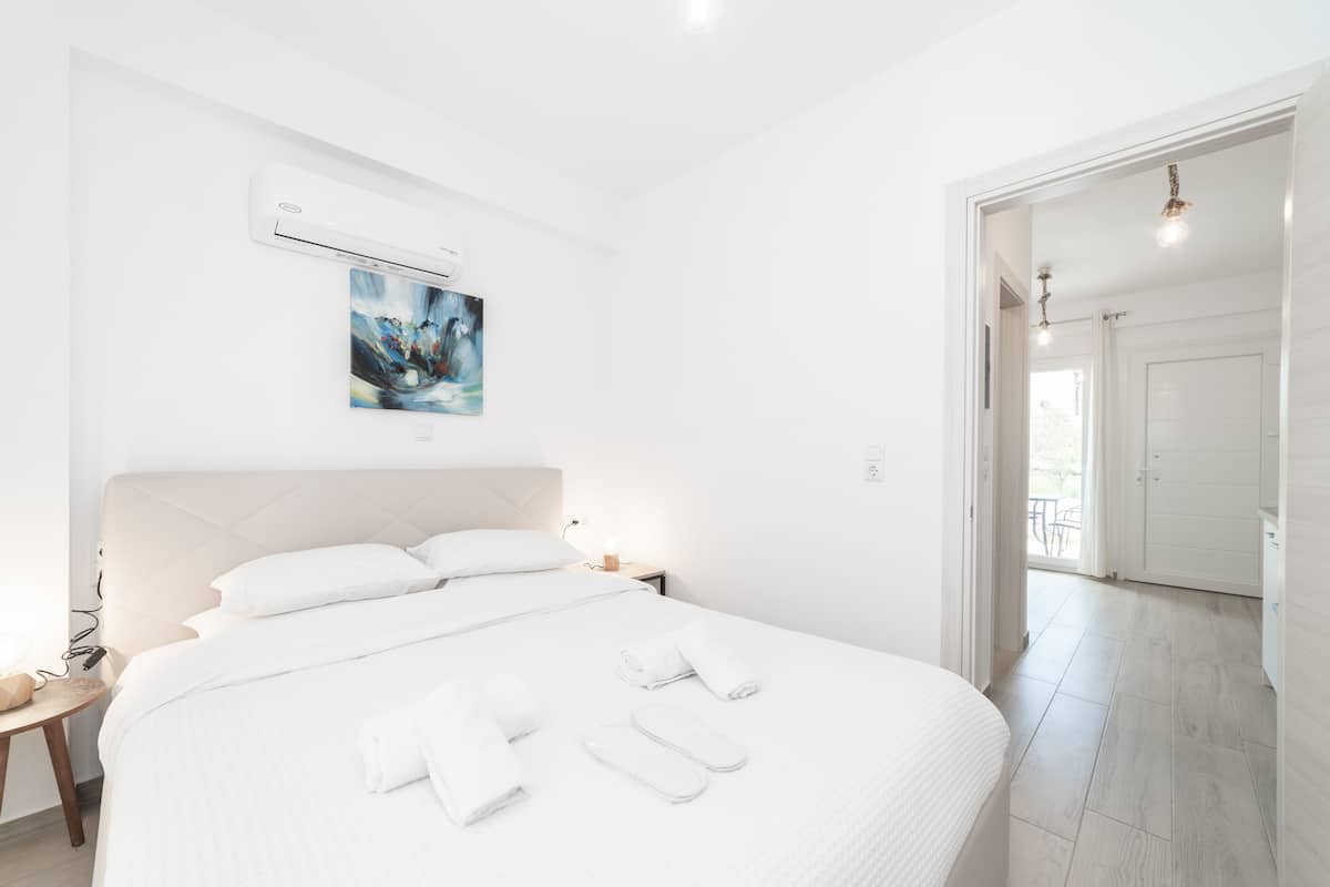 A modern hotel room with a large bed, white linens, a painting on the wall, and a view to the outside.