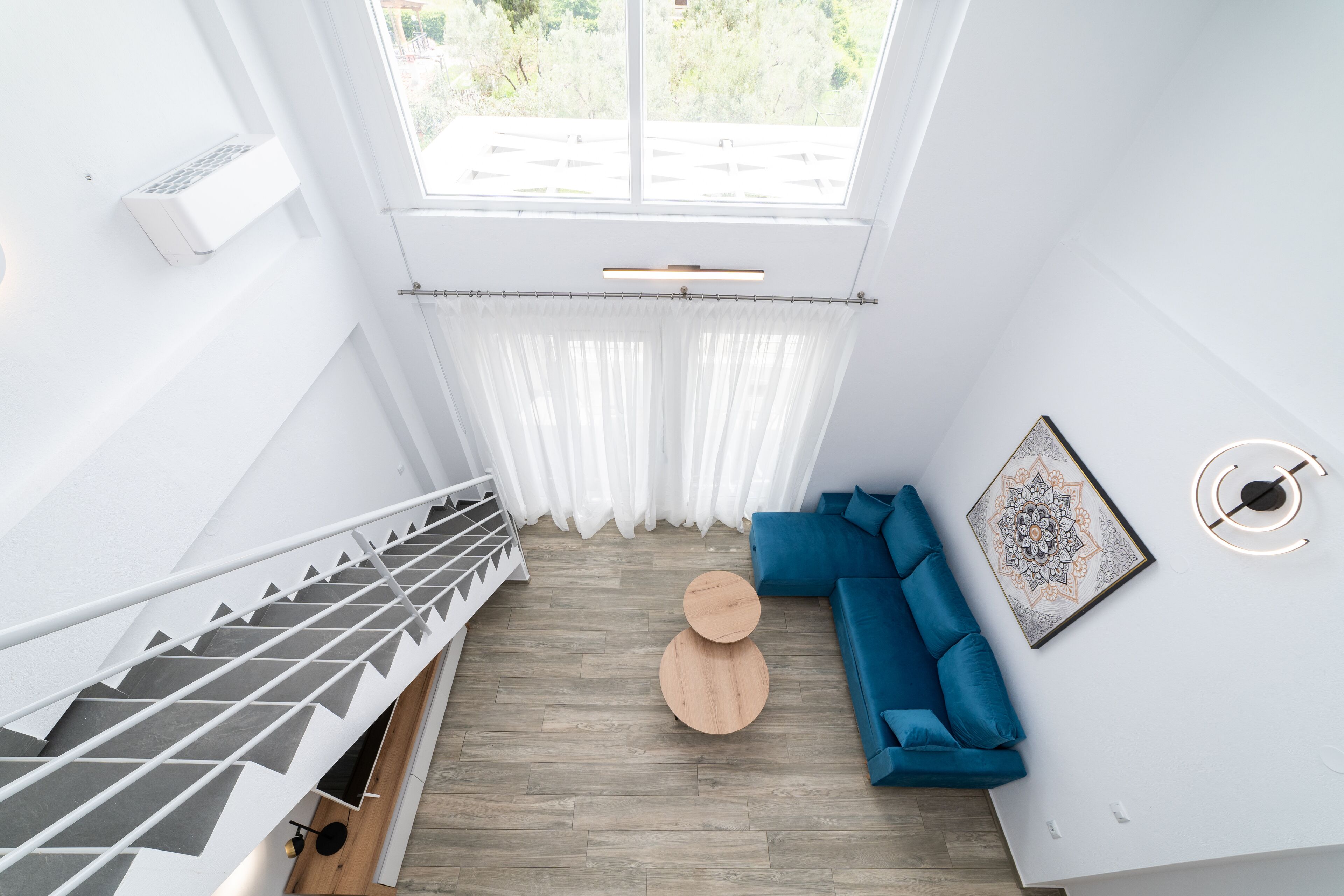 Un intérieur moderne avec un escalier, un canapé bleu, une table basse ronde en bois et une grande fenêtre avec des rideaux.