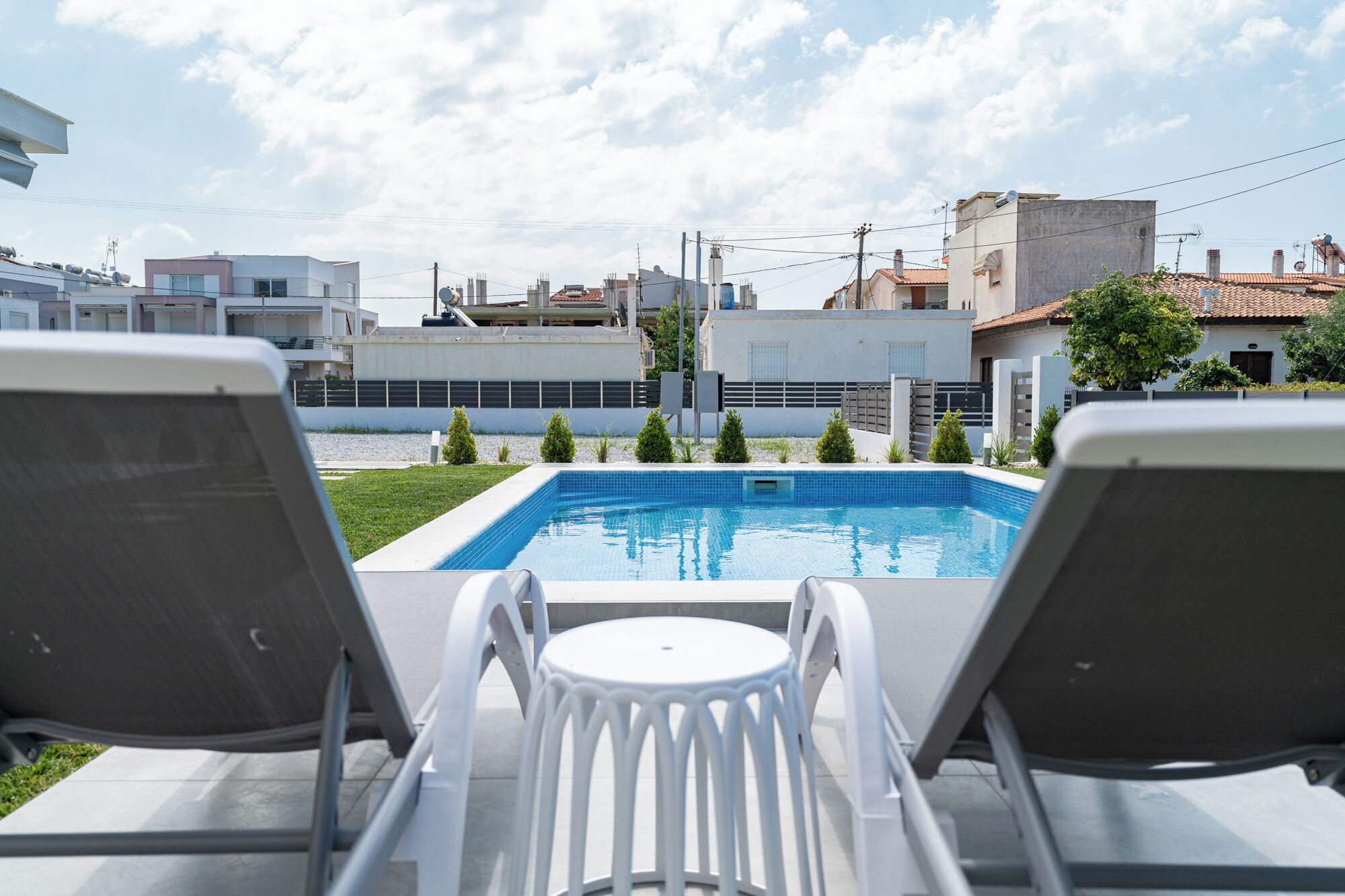 A swimming pool with two lounge chairs facing it, surrounded by a low fence and small trees.