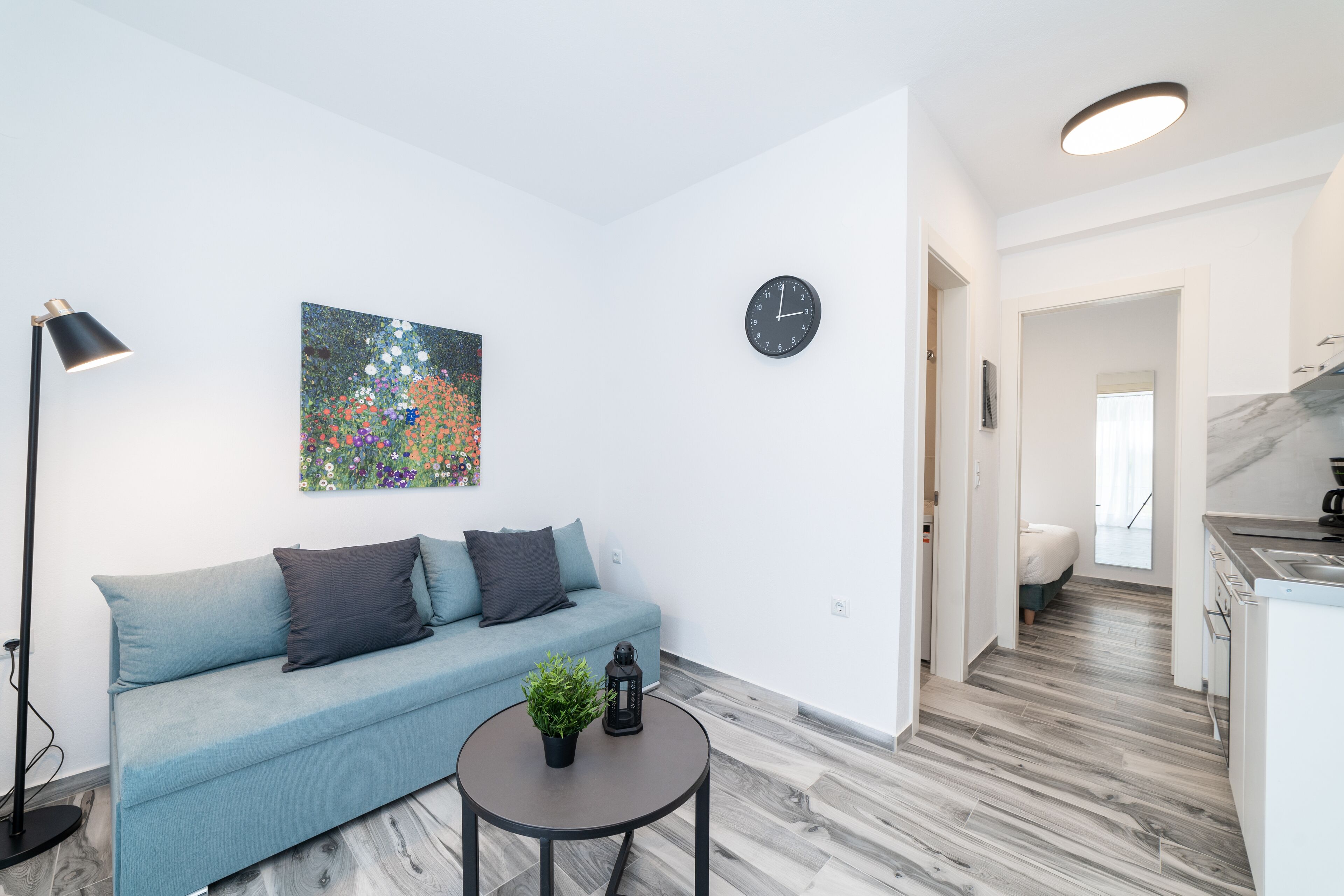 Una sala de estar moderna con un sofá azul claro, una mesa redonda con una planta, un reloj negro y un cuadro en la pared.
