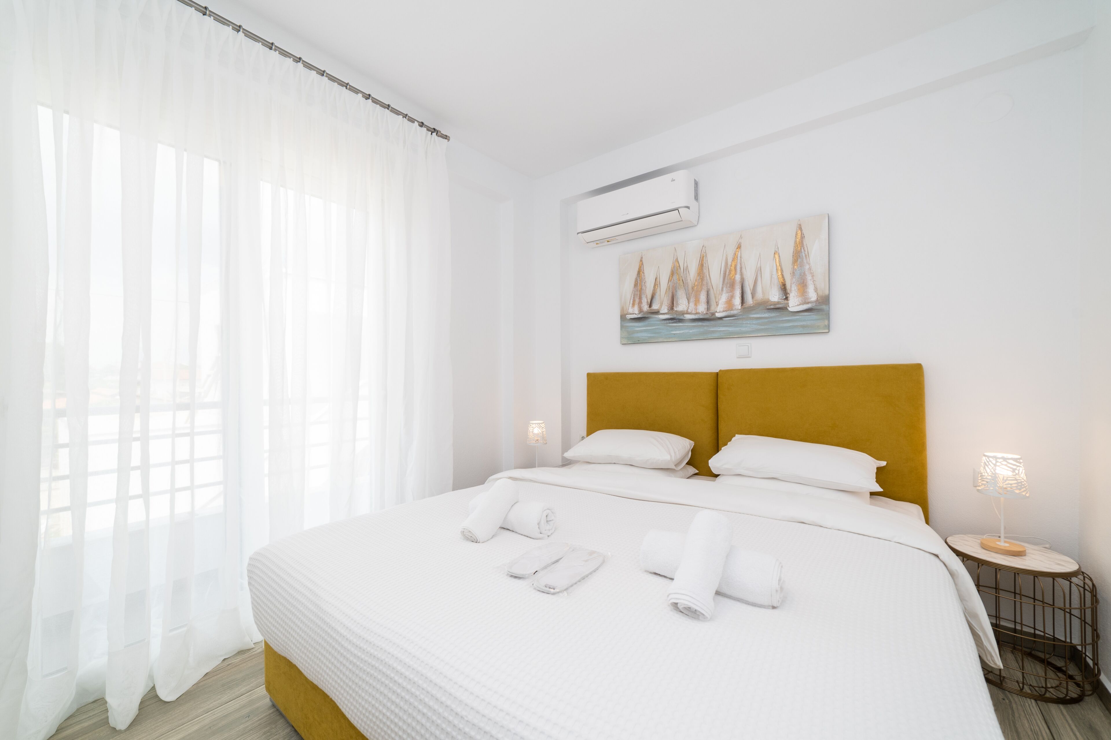 A hotel room with a large bed, white bedding, a headboard with a yellow accent, a painting of sailboats, an air conditioning unit, and a bedside lamp.