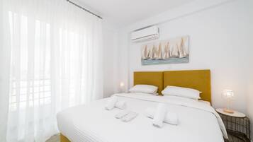 A hotel room with a large bed, white bedding, a headboard with a yellow accent, a painting of sailboats, an air conditioning unit, and a bedside lamp.