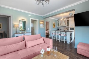 A living room with a pink sofa, a coffee table with a bottle and glasses, and a TV in the background.
