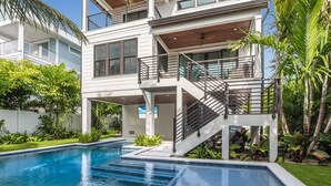 A modern two-story house with a swimming pool, a covered outdoor area, and lush greenery. - Luxe Island Escape by Beach Boutique Rentals (Anna Maria)