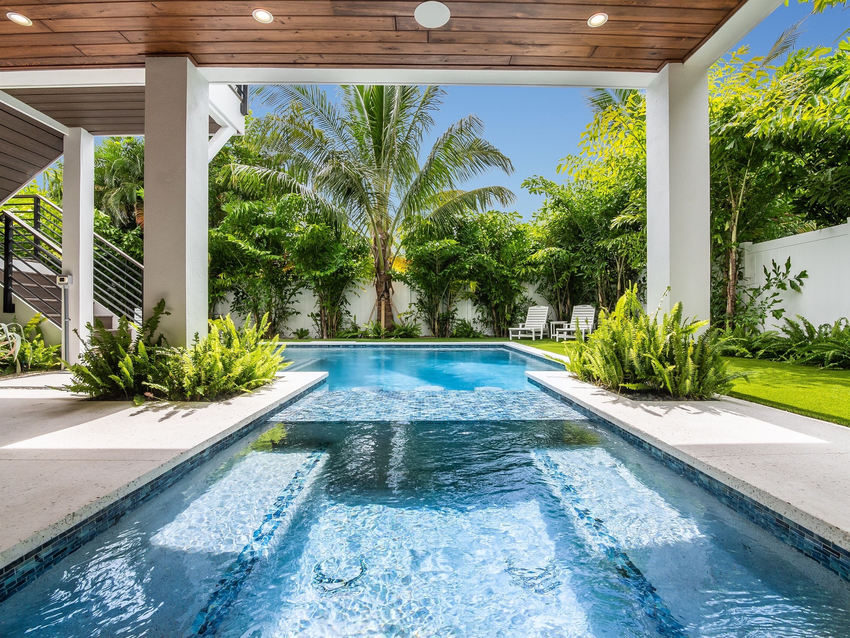 Piscina retangular com águas azuis cristalinas, rodeada por vegetação exuberante e colunas brancas.