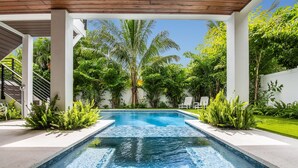 A rectangular swimming pool with clear blue water, surrounded by lush greenery and white columns. - Luxe Island Escape by Beach Boutique Rentals (Anna Maria)
