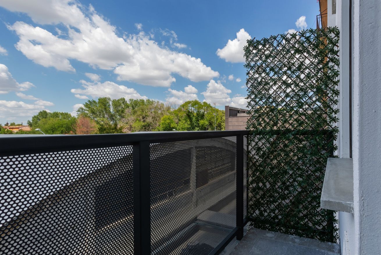 Een balkon met een zwart metalen hekwerk, een gaaswerk en een helderblauwe lucht met wolken.