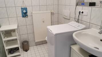 A small, clean bathroom with a white sink, a washing machine, and a shelf with toiletries.