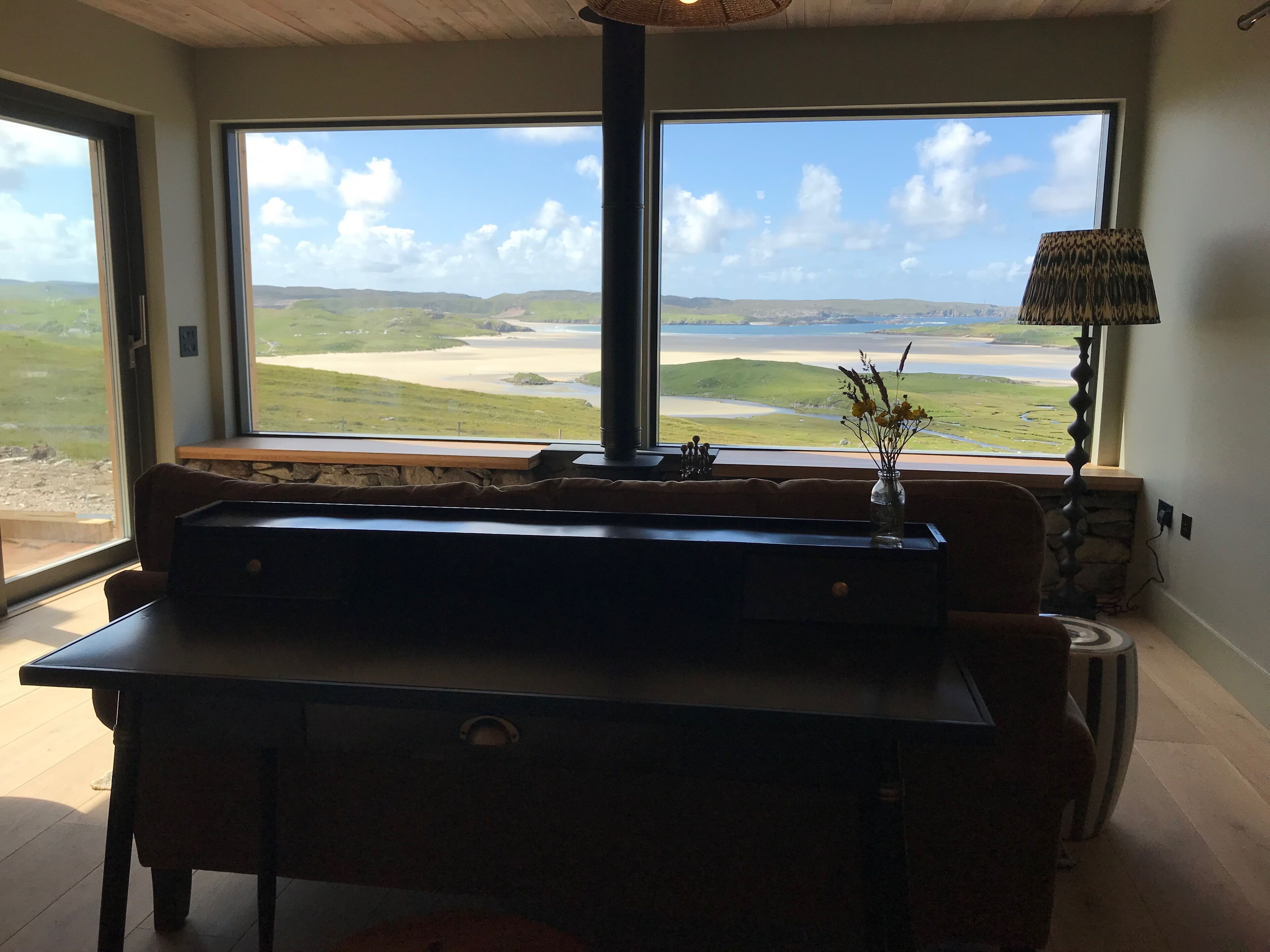 A room with a large window offering a view of a coastal landscape, a wooden table, a vase with flowers, and a lamp.