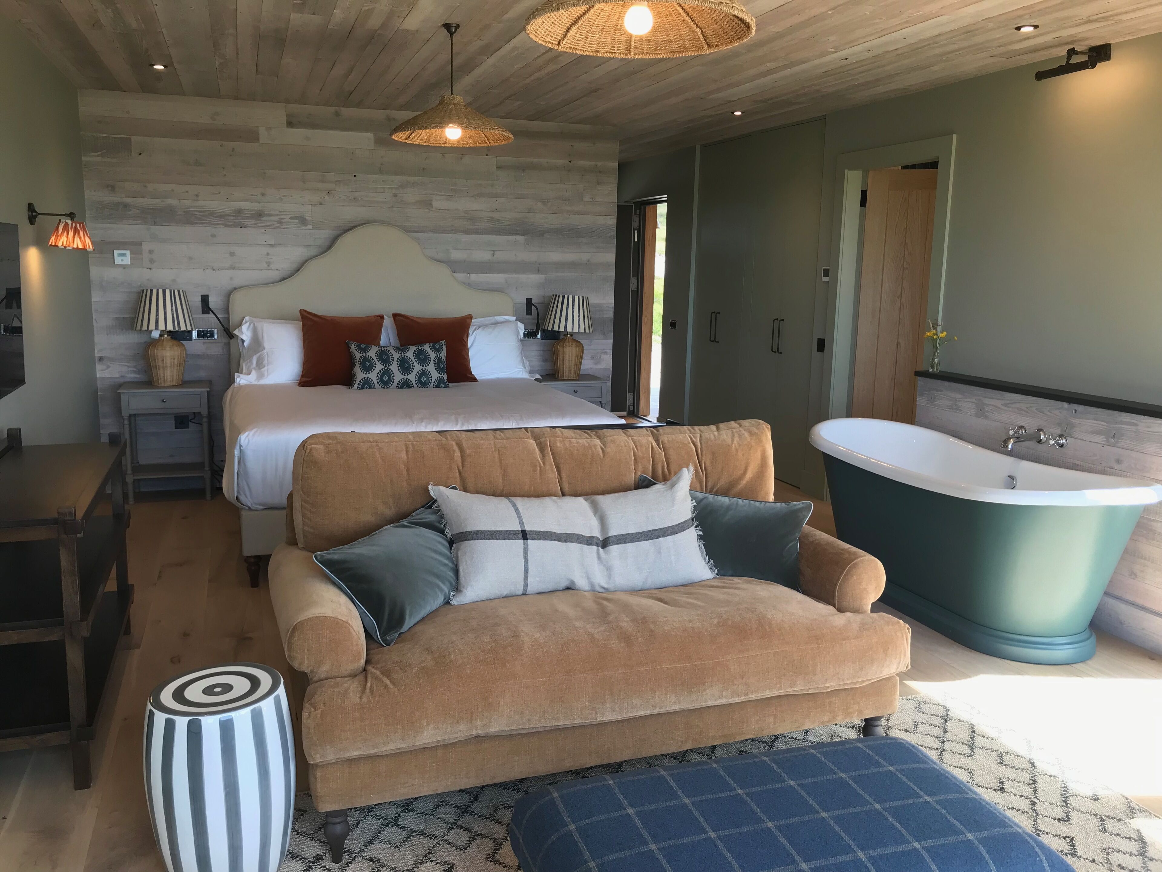 A bedroom with a large bed, a sofa, a side table, a lamp, a hanging light, and a bathtub.