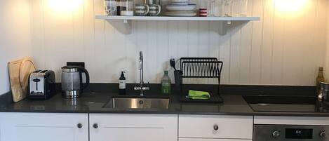 Une cuisine avec des armoires blanches, un plan de travail noir et un plafond en bois. On y trouve deux luminaires fixés au mur, une machine à café, un grille-pain et un égouttoir à vaisselle.