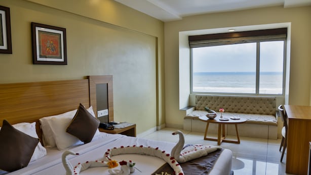 A hotel room with a bed, a desk, a chair, a window with a view of the sea, and a framed picture on the wall.
