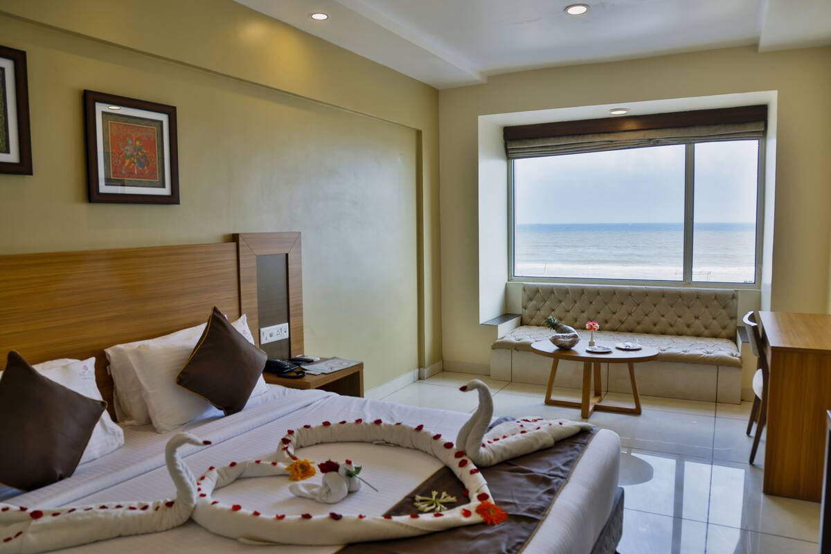 A hotel room with a bed, a desk, a chair, a window with a view of the sea, and a framed picture on the wall.