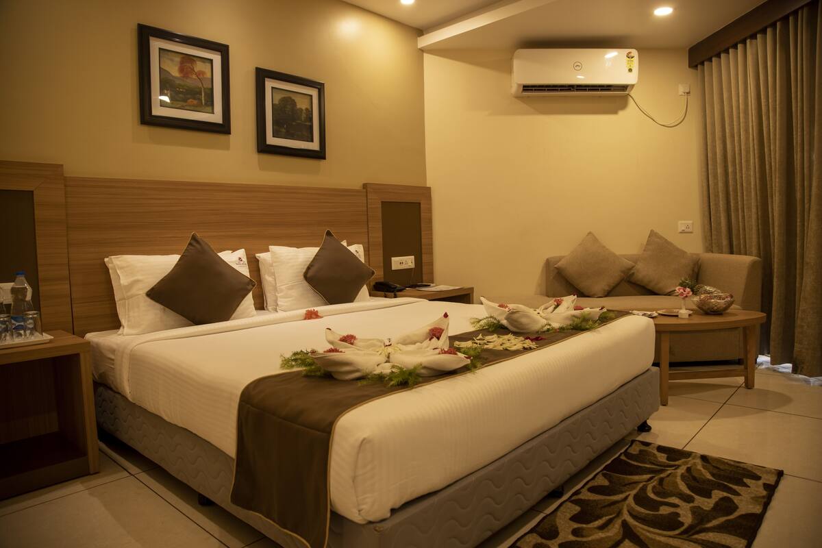 A hotel room with a large bed, a sofa, a nightstand, and an air conditioning unit.