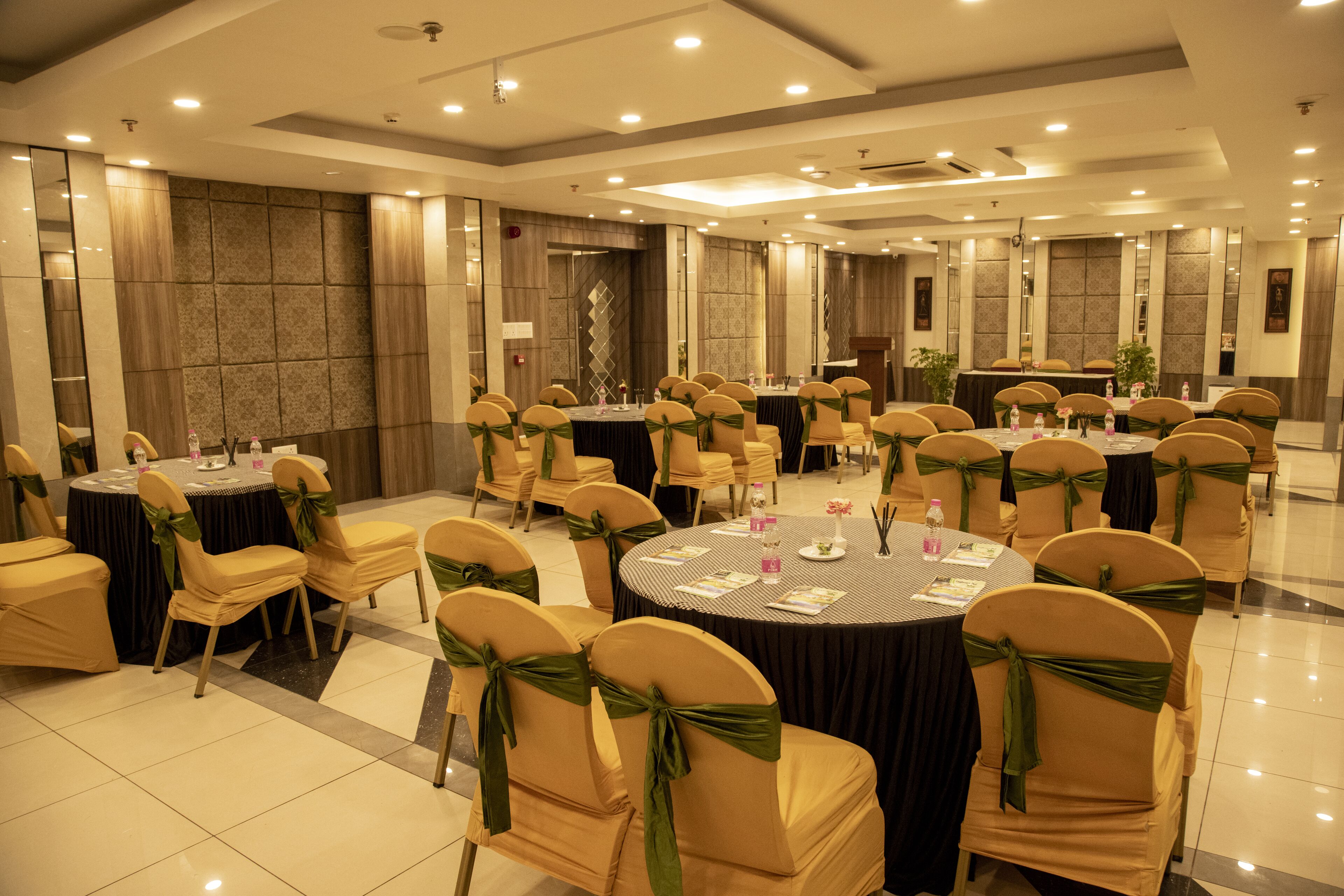 A conference room with multiple round tables, each surrounded by chairs with green sashes, and a stage area with a podium.