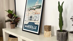 A white bench with a framed picture of Marseille, a potted plant, a cactus, and a glass jar.
