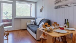 A cozy room with a sofa, a dining table set with plates and glasses, and a window with a view of greenery. - Les Lauzes D25 by Interhome (Les Menuires)