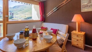 Een gezellige kamer met een houten eettafel gedekt voor twee personen, een houten stoel, een klein houten dressoir, een raam met uitzicht op bergen en een schilderij aan de muur.