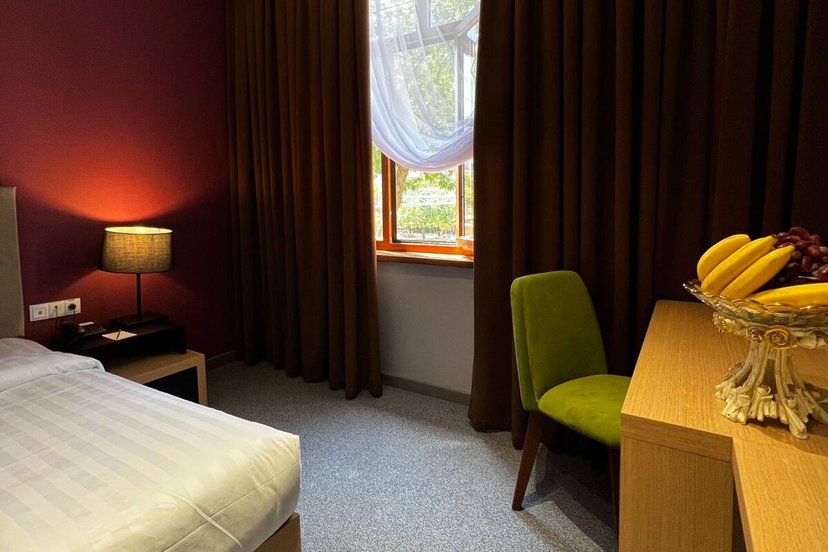 A hotel room with a bed, a desk with a fruit bowl, a chair, a lamp, and a window with curtains.