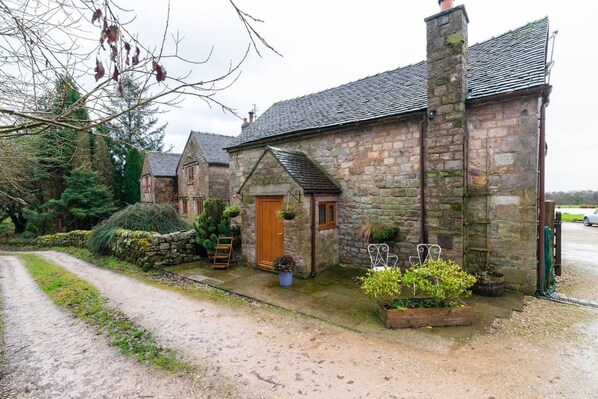 A stone cottage with a dirt path leading to the entrance, surrounded by greenery and a stone wall.