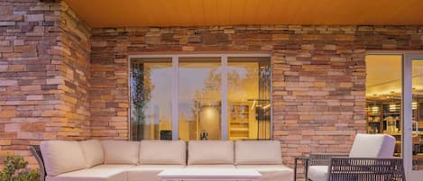 Une terrasse extérieure avec un mur en pierre, un canapé beige, une table blanche et des chaises en osier.