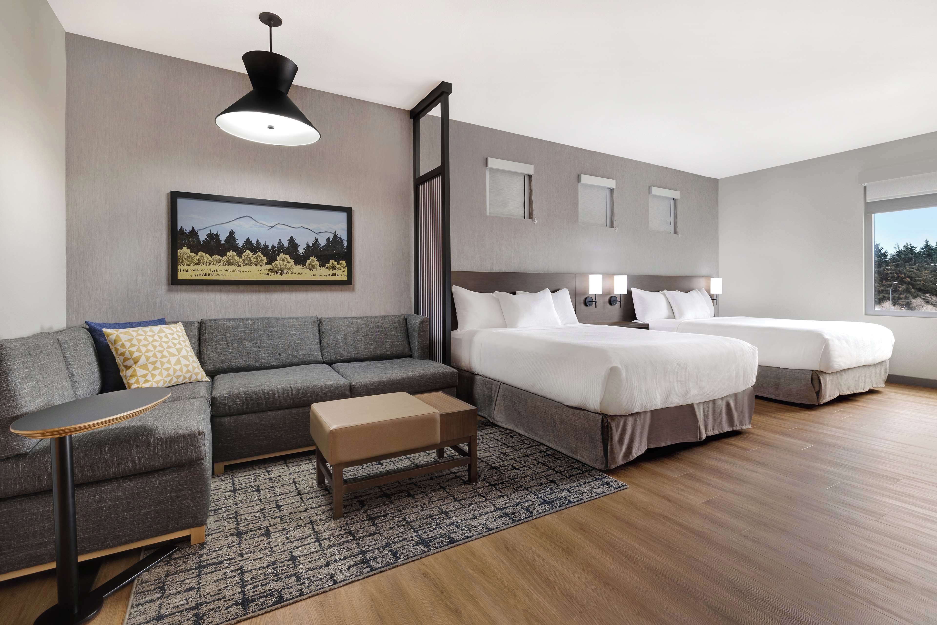 A modern hotel room with a grey sofa, a small table, a bed with two pillows, and a large window.