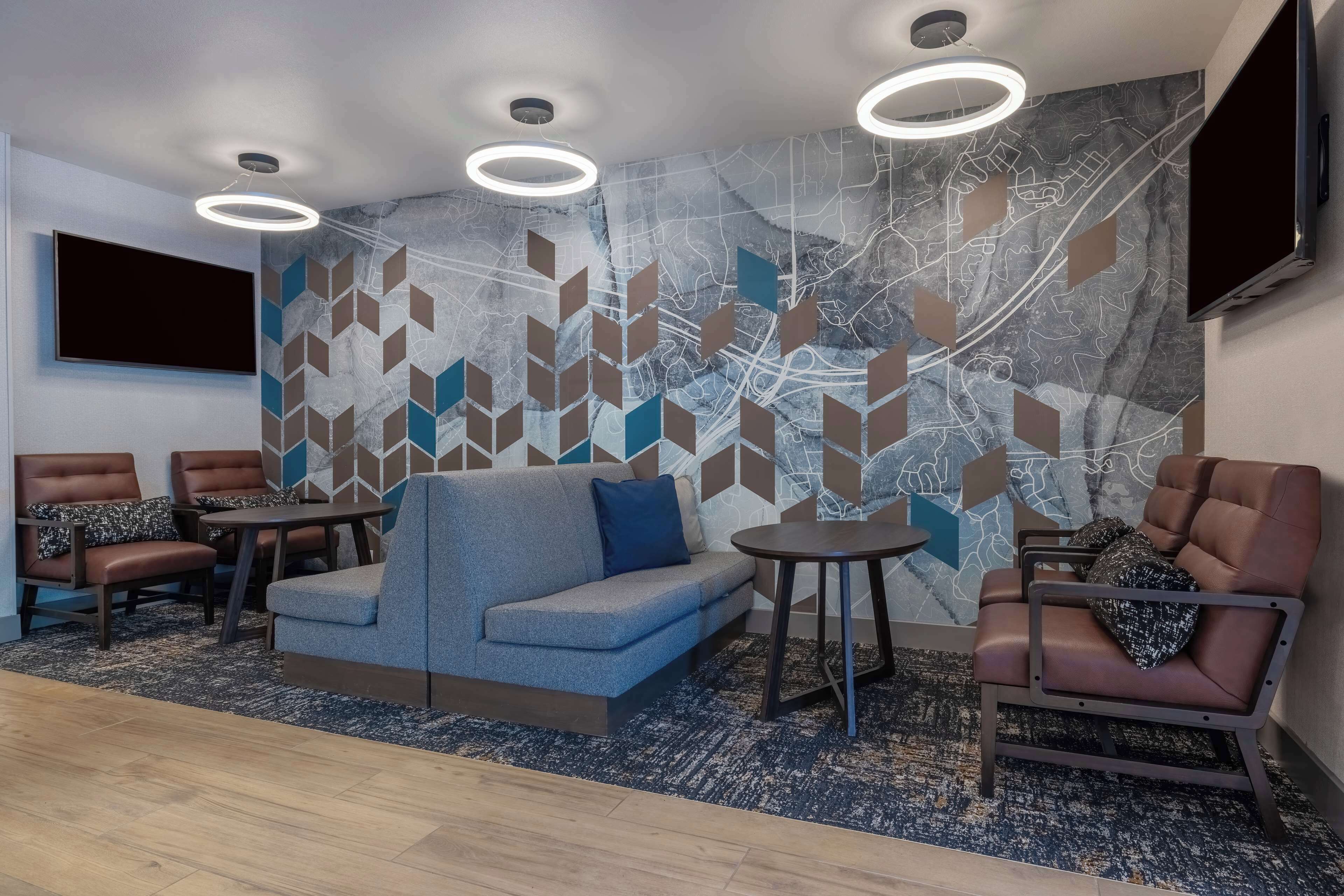A modern lounge area with a sofa, armchairs, and tables, featuring a geometric patterned wall and a flat-screen TV.