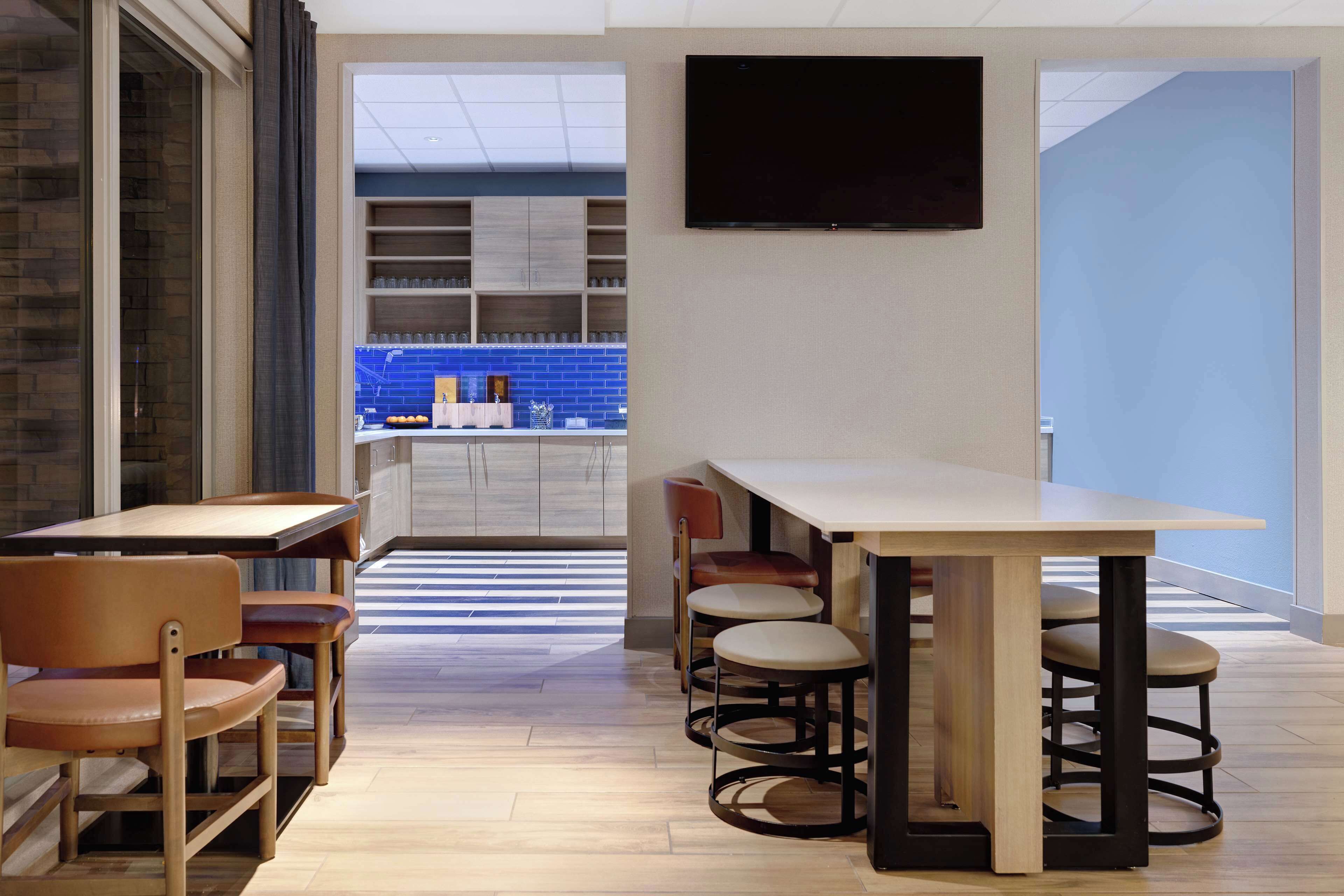 A modern dining area with a long table and multiple chairs, a kitchen in the background, and a flat-screen TV mounted on the wall.
