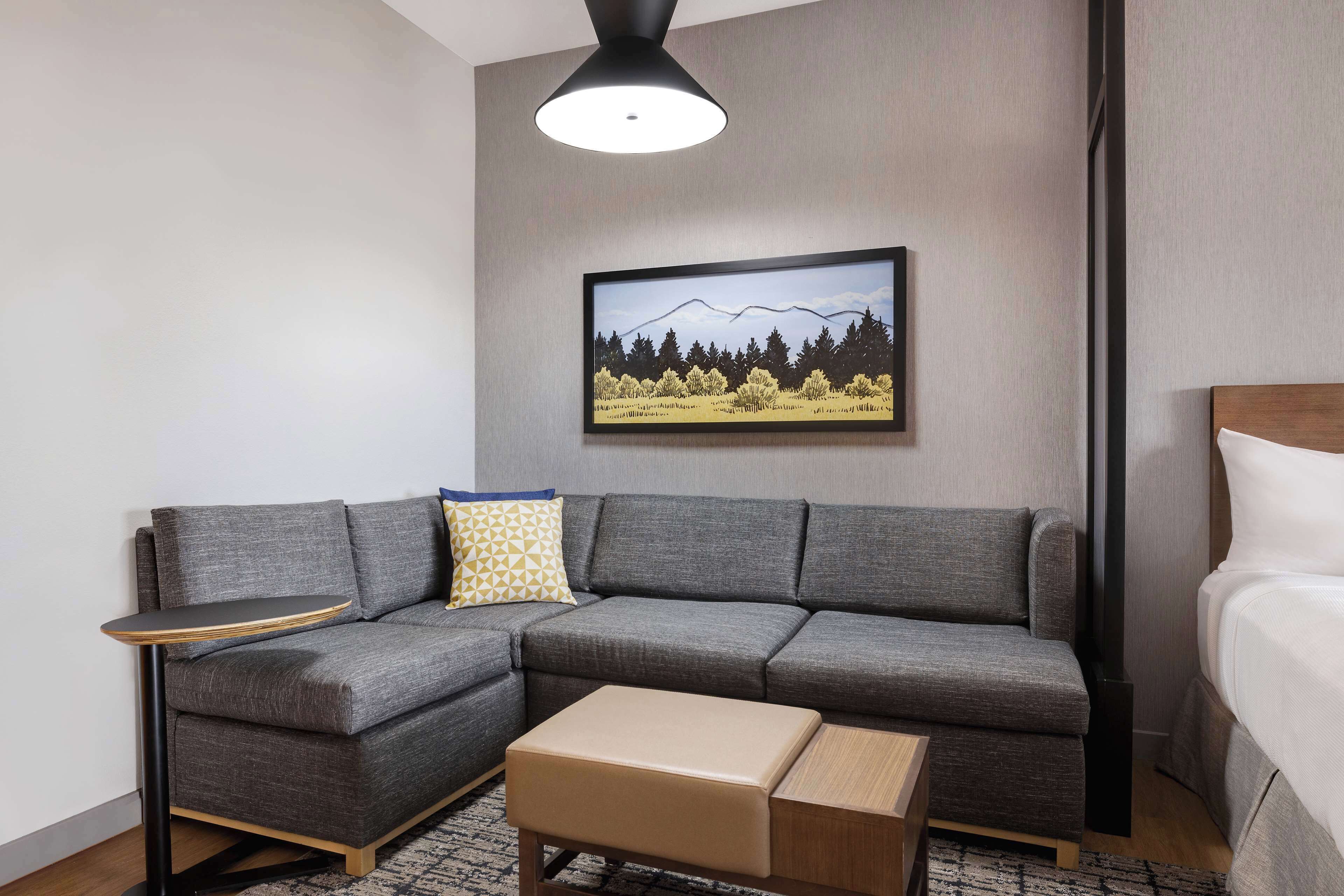 A modern hotel room with a grey sectional sofa, a small wooden side table, a beige ottoman, a framed landscape picture on the wall, and a bed with white linens.