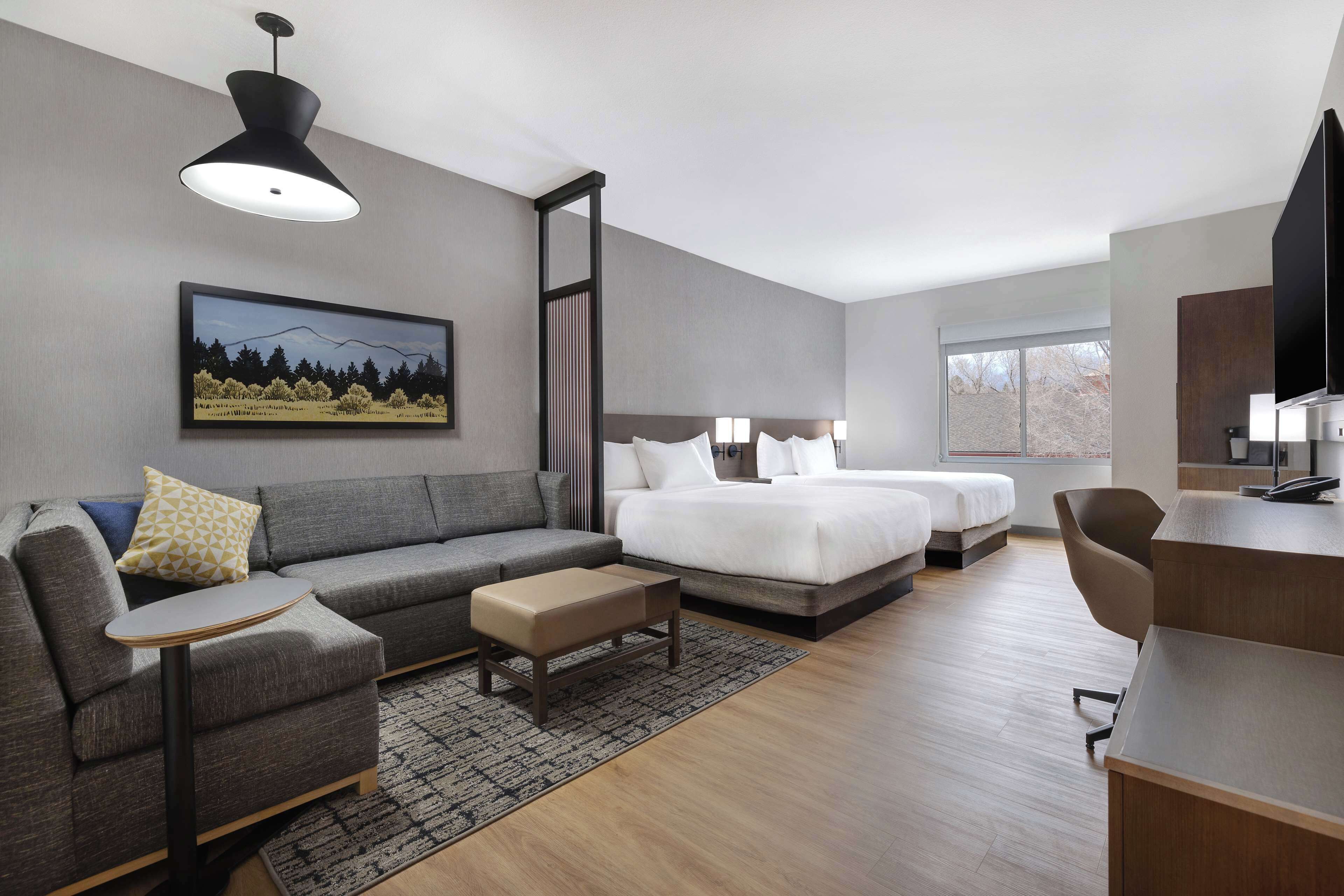 A modern hotel room with a grey sofa, a small table, a bed with two pillows, a desk with a chair, and a large window.