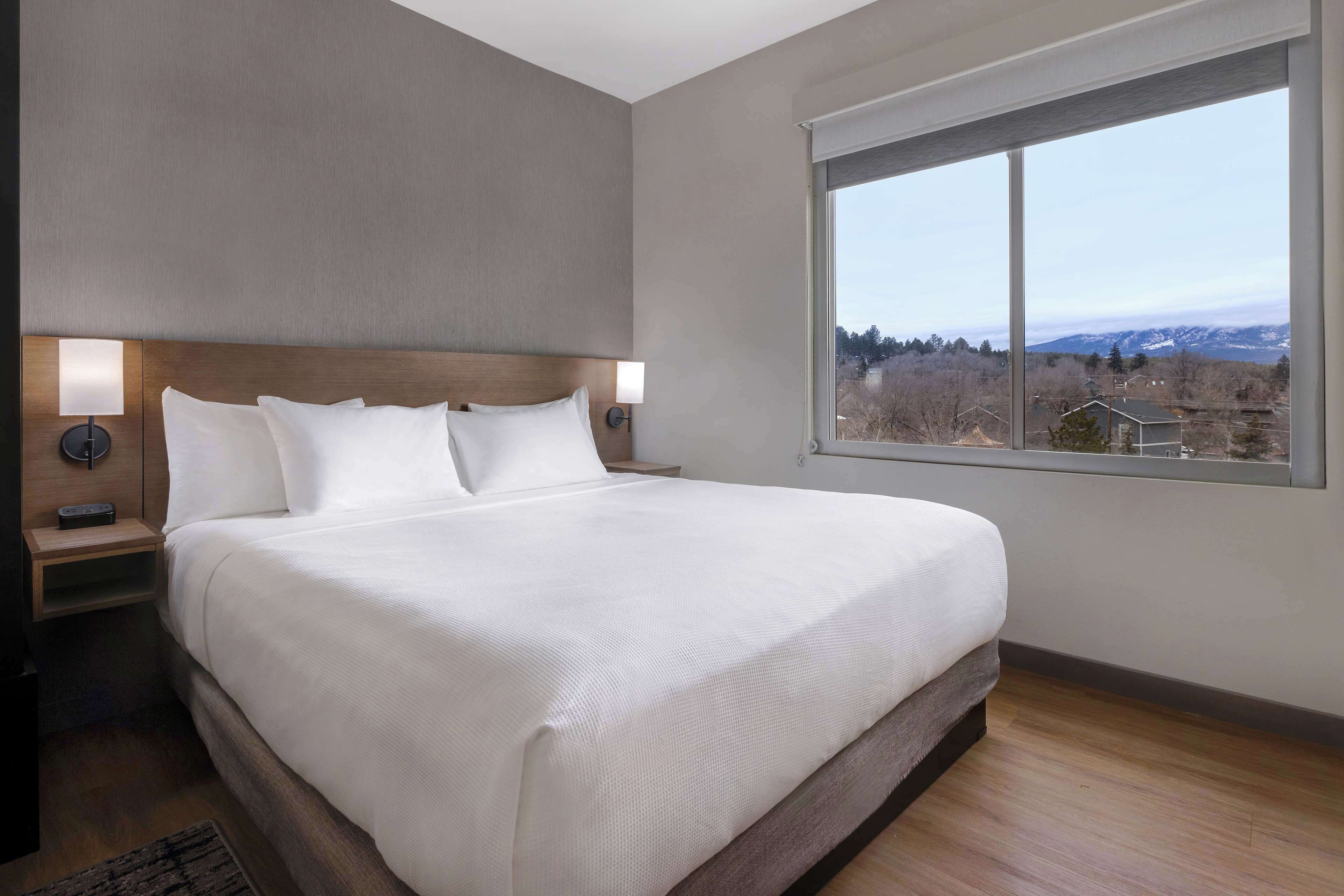 A modern hotel room with a large bed, a nightstand, a lamp, and a view of a mountainous landscape through a large window.