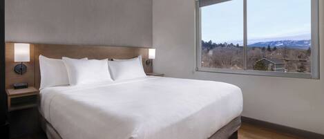 A modern hotel room with a large bed, a nightstand, a lamp, and a view of a mountainous landscape through a large window.