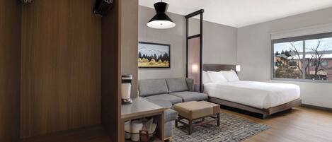 A modern bedroom with a bed, a gray sofa, a wooden wardrobe, a desk, and a large window.