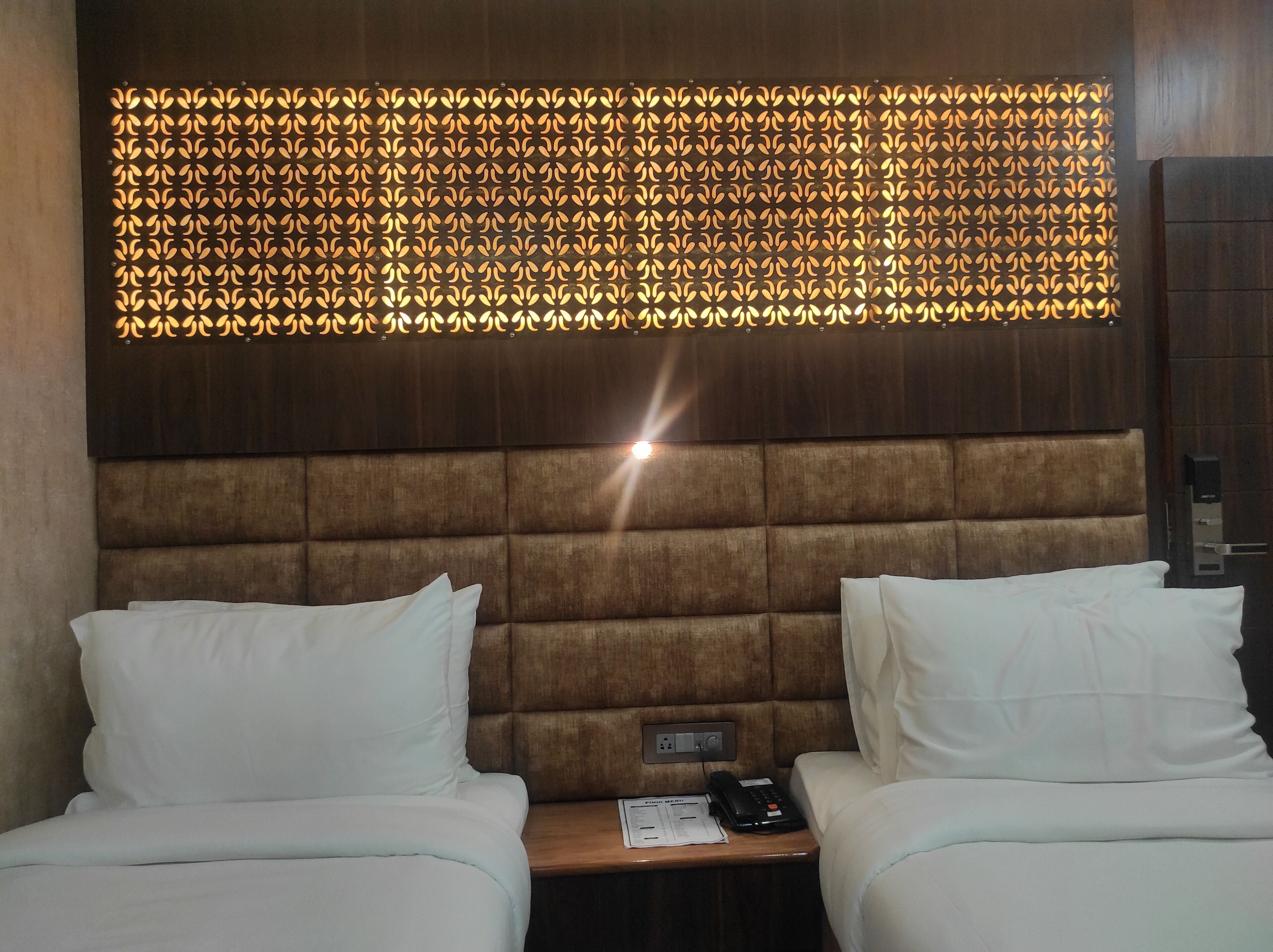 A hotel room with a tufted headboard, two beds with white linens, a wall-mounted light fixture, and a bedside table with a phone and papers.