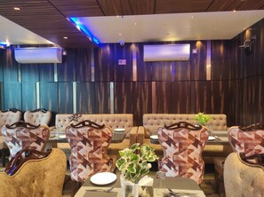 A restaurant with a modern interior, featuring wooden panel walls, air conditioning units, and a mix of patterned and tufted seating.