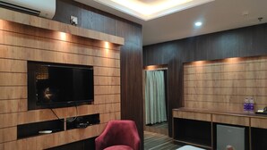 Una habitación de hotel moderna con televisión de pantalla plana montada en una pared de paneles de madera, un sillón rojo, una mesita redonda y un minibar.