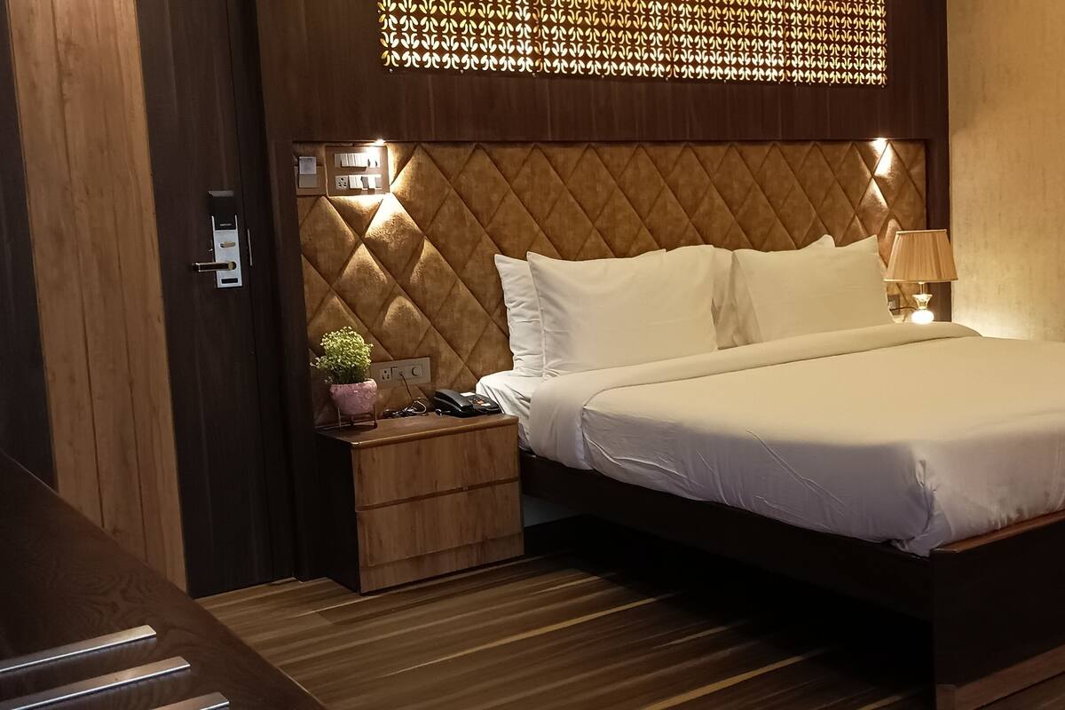 A hotel room with a large bed, wooden headboard, bedside table with a lamp, and a small plant.