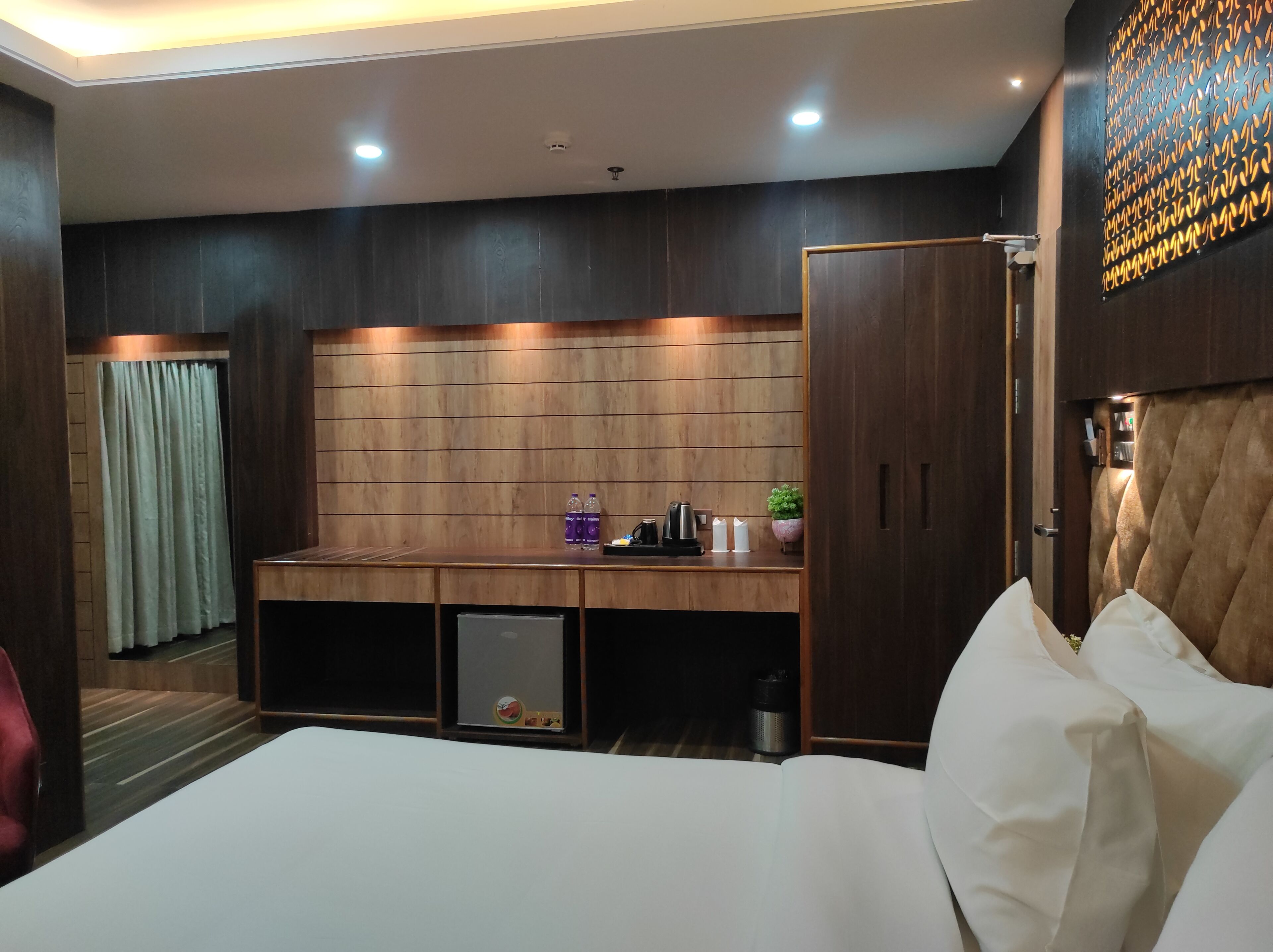 Une chambre d’hôtel avec un grand lit, un bureau en bois, une machine à café, des bouteilles et une petite plante.