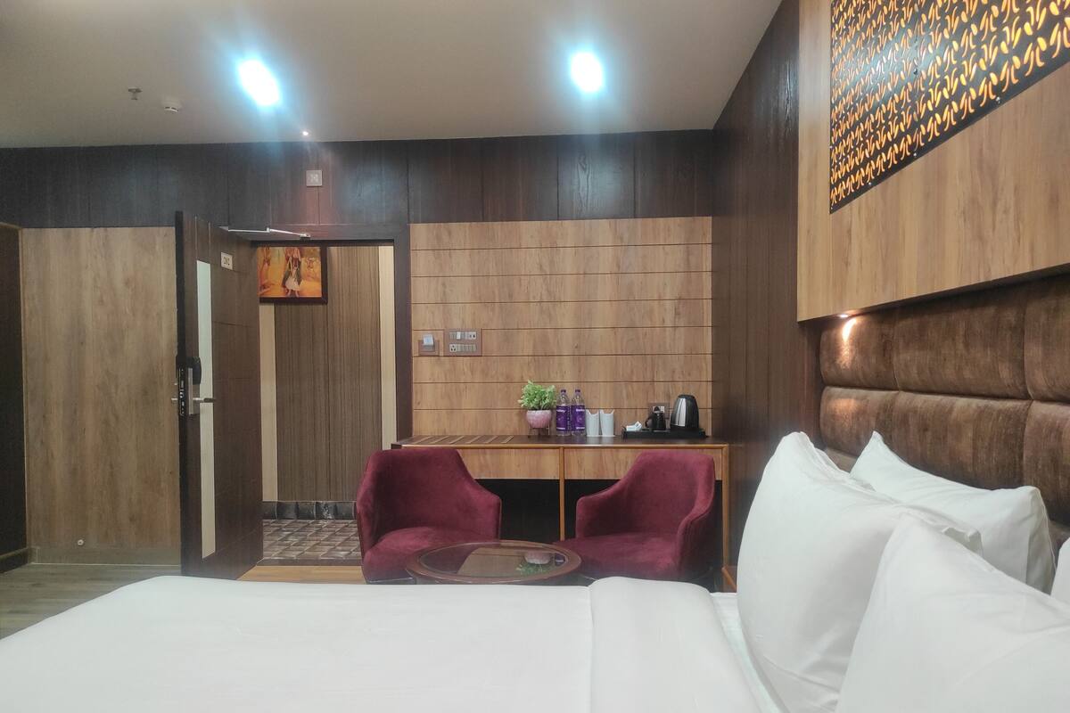 A hotel room with a bed, two red chairs, a small table, a desk with a plant, and a wall with a patterned design.