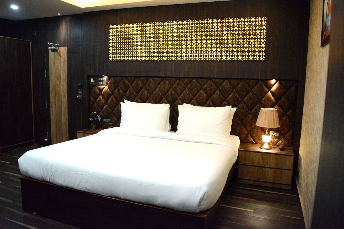 A modern hotel room with a large bed, wooden headboard, a wall-mounted light fixture, and a bedside table with a lamp.