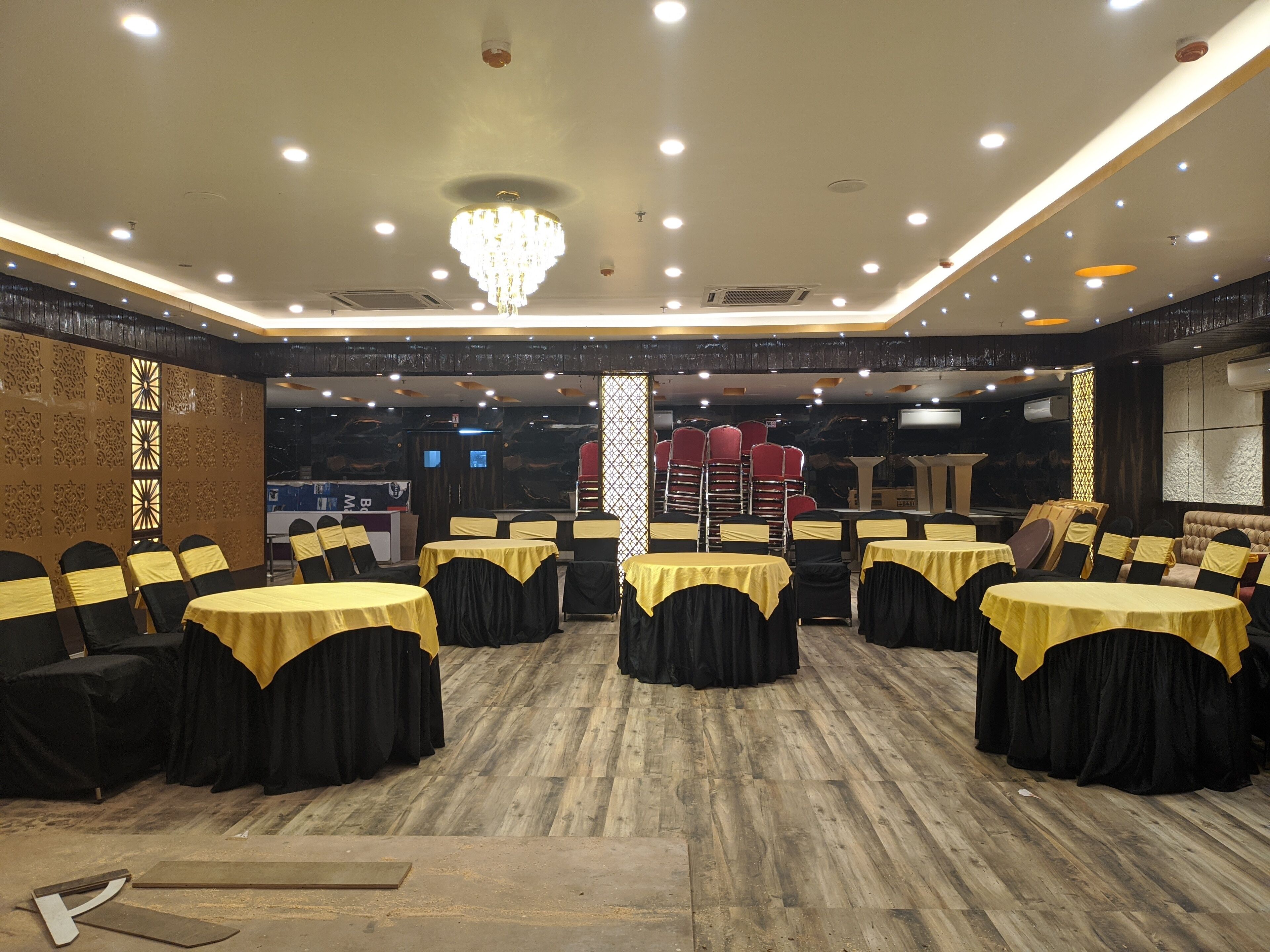 A banquet hall with round tables covered in yellow tablecloths and black chairs, wooden flooring, and a chandelier.