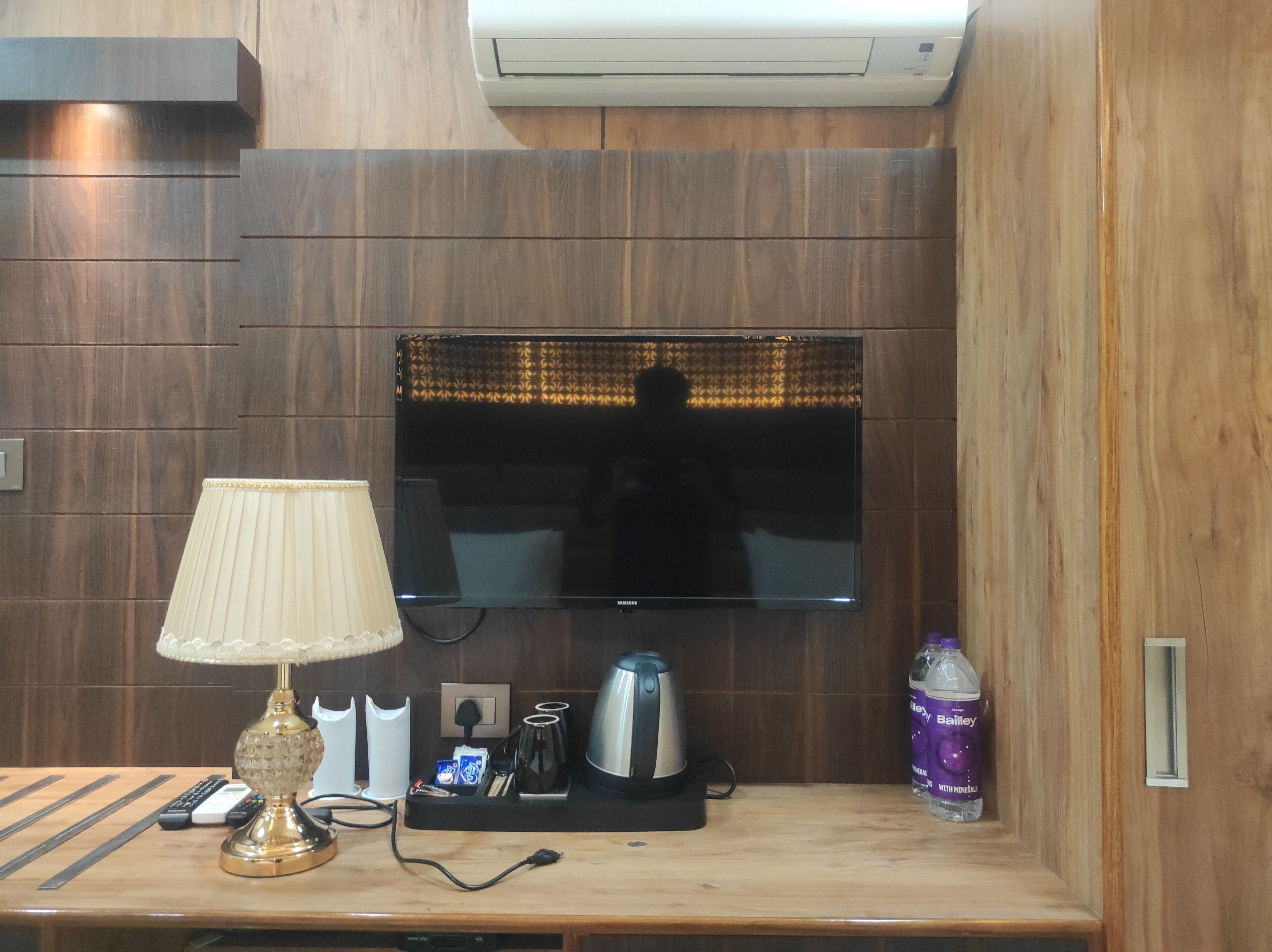 A hotel room with a flat-screen TV mounted on the wall, a bedside table with a lamp, a coffee maker, a bottle of Bailey's, and a remote control.