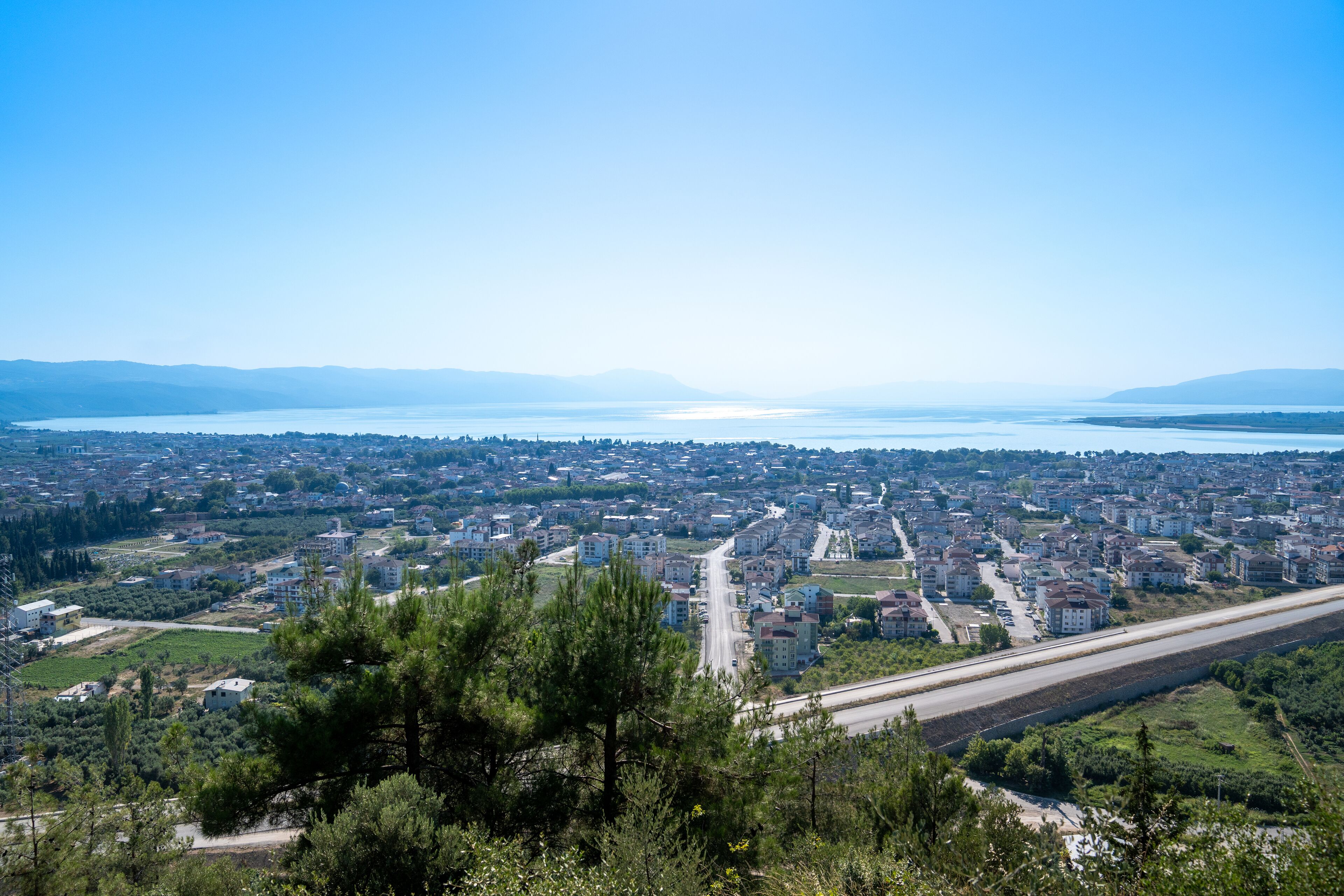 Vista panorámica de una ciudad costera con una carretera despejada que conduce al pueblo, rodeada de vegetación y edificios residenciales.
