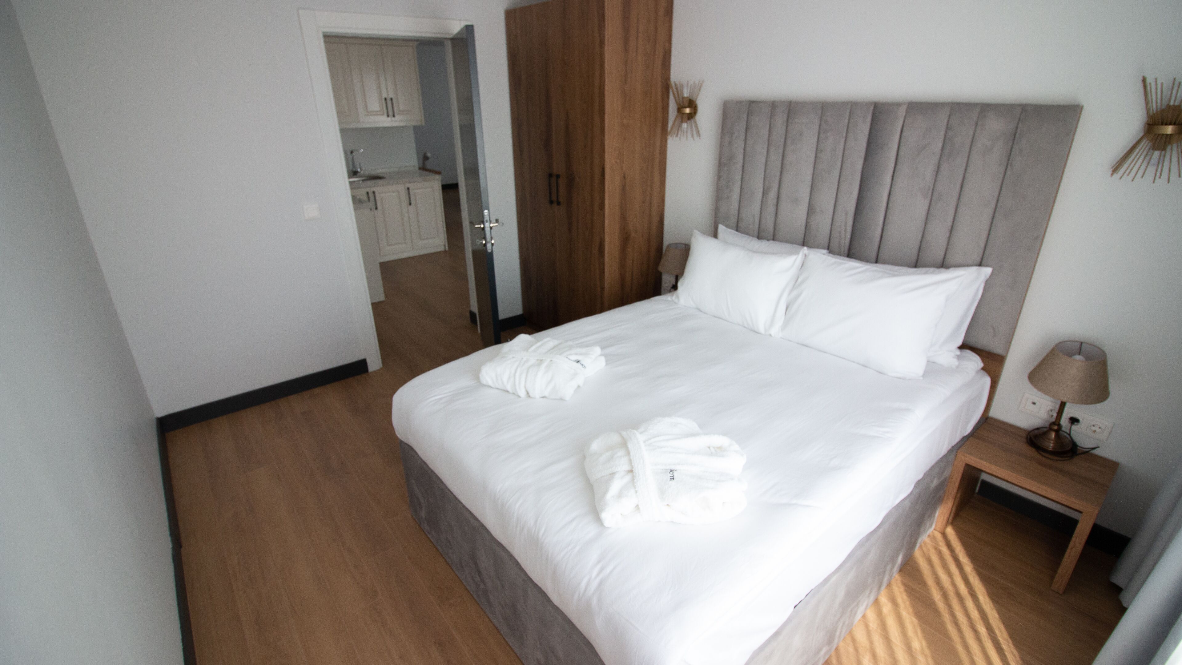 A hotel room with a large bed, a wooden wardrobe, a bedside table with a lamp, and a view of the kitchen.