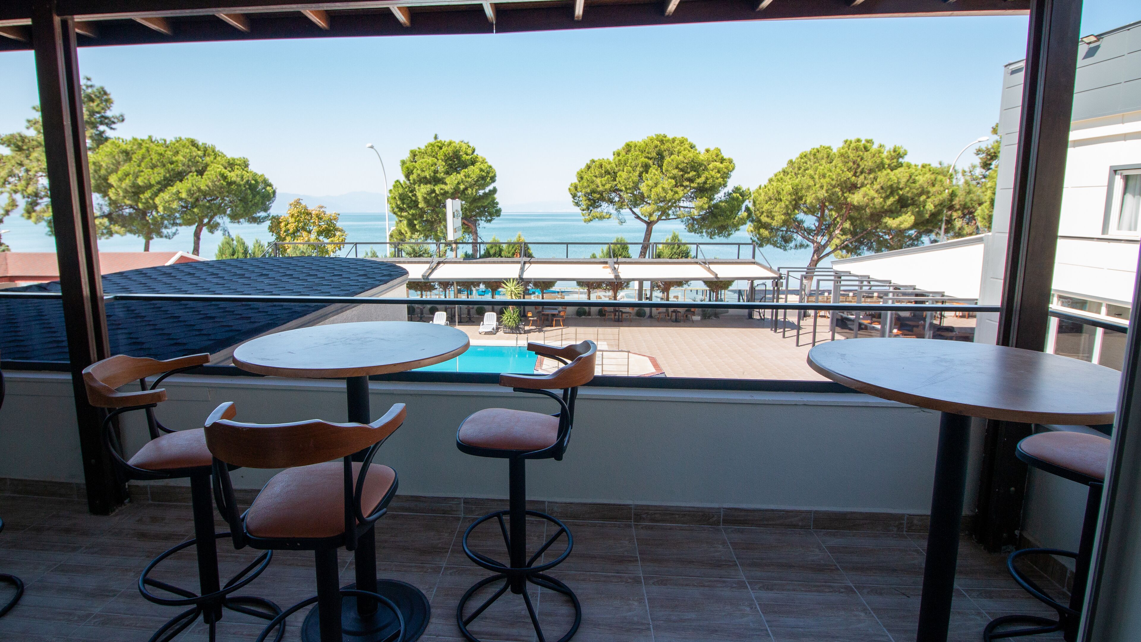 A terrace with two tables and chairs, overlooking a pool and outdoor seating area with trees and a view of the sea.